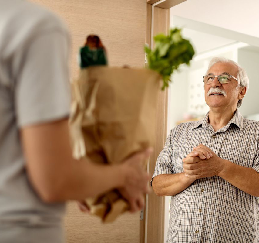A man standing at the doorway is about to receive a paper bag of groceries